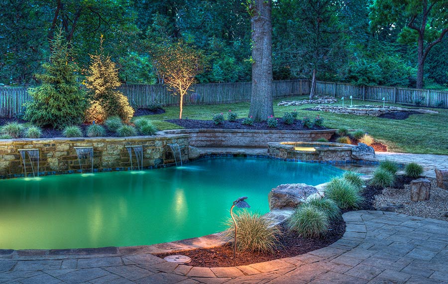A beautifully lit backyard pool resort by Backyard by Design
